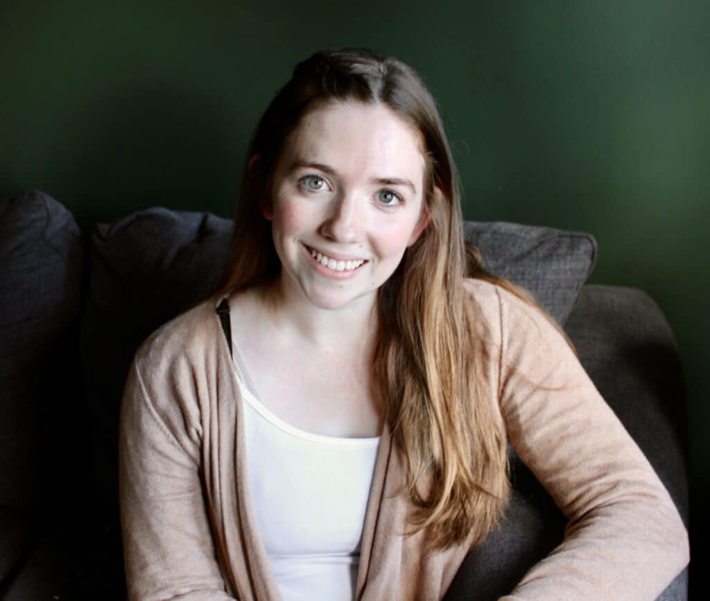 Abigael Davis, LCSW, Trauma Therapist with light brown hair, smiling sitting on a gray couch wearing a white shirt and cream cardigan.